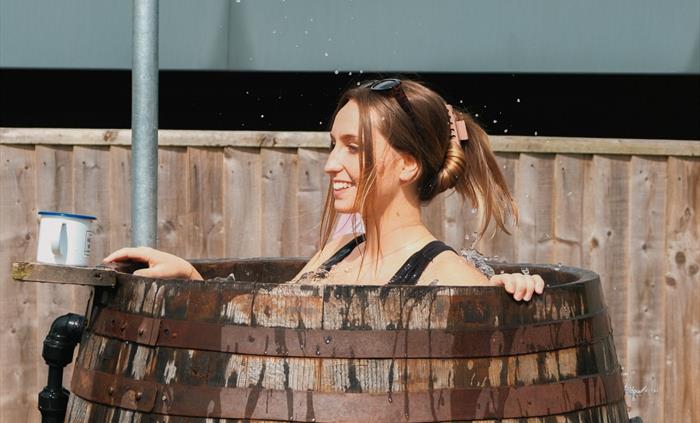 Young woman in a Halen Môn Wild Seaweed Bath, splashing in the barrel with a Halen Môn drink to hand, in the sunshine